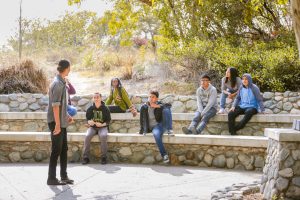 A picture of students in an outdoor park seated and facing one student who is standing and talking to them.